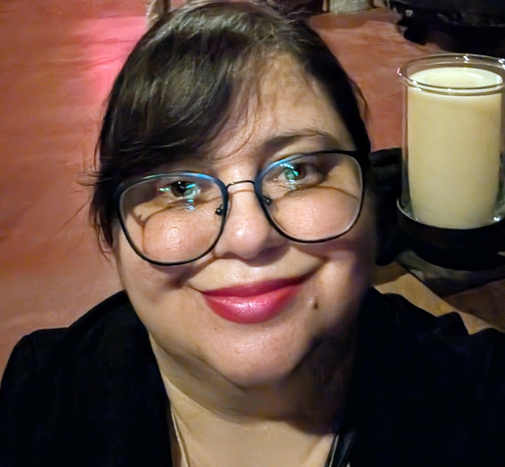 A close-up selfie of a woman with dark hair and large glasses, smiling gently. She has a light pink lipstick on and is sitting in a dimly lit environment with a candle in the background.