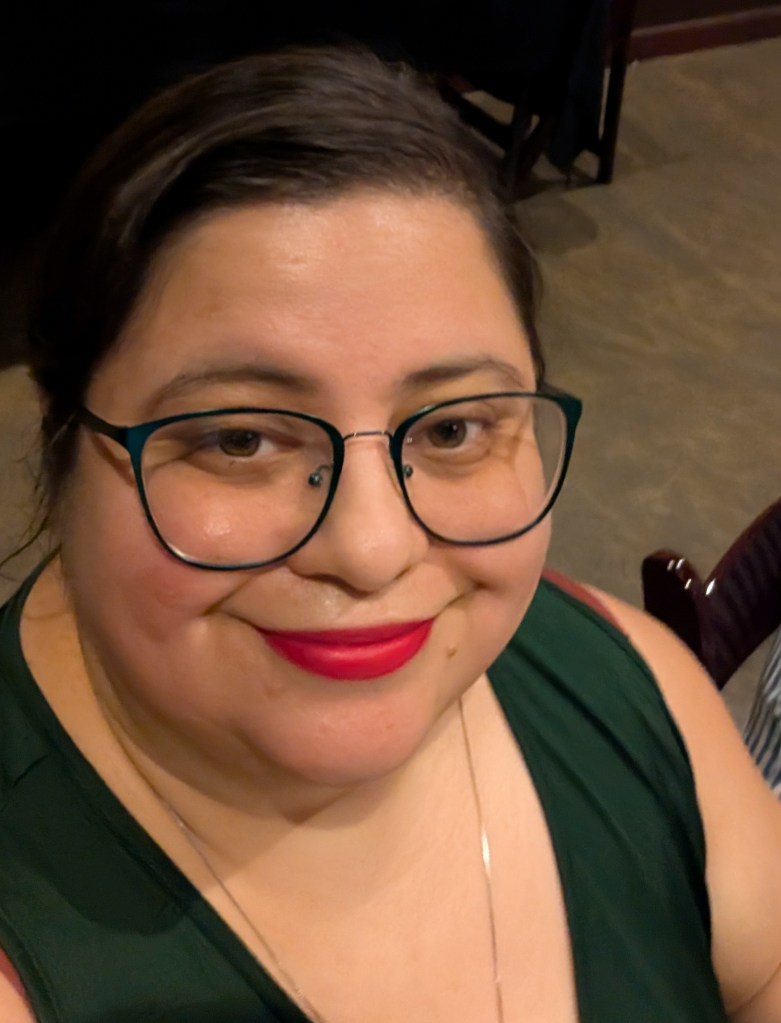 A smiling woman with glasses and bright red lipstick, wearing a green top, indoors.